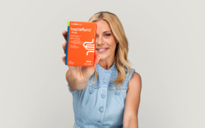 Woman smiling and presenting an orange Bactflora Plus probiotic box toward the camera.