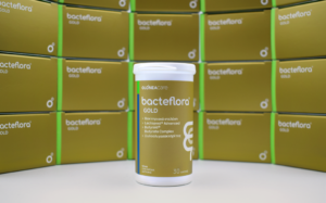 Probiotic supplement bottle labeled Bacteflora GOLD centered on a white surface, with gold packaging boxes stacked behind.