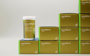 White bottle labeled 'bacteflora GOLD' sits on a stack of gold-and-green boxes with matching branding in a clean studio setting.