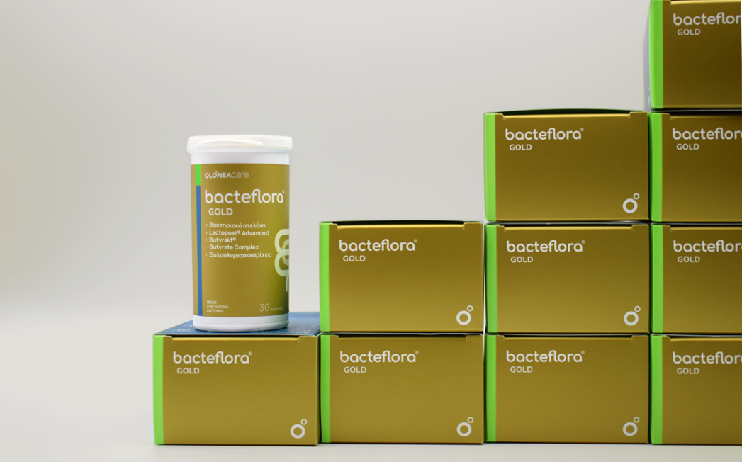 White bottle labeled 'bacteflora GOLD' sits on a stack of gold-and-green boxes with matching branding in a clean studio setting.