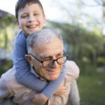 Ένας παππούς με το εγγόνι του, το οποίο έχει στους ώμους του και φαίνονται να απολαμβάνουν χαρούμενες στιγμές στον κήπο του σπιτιού τους.