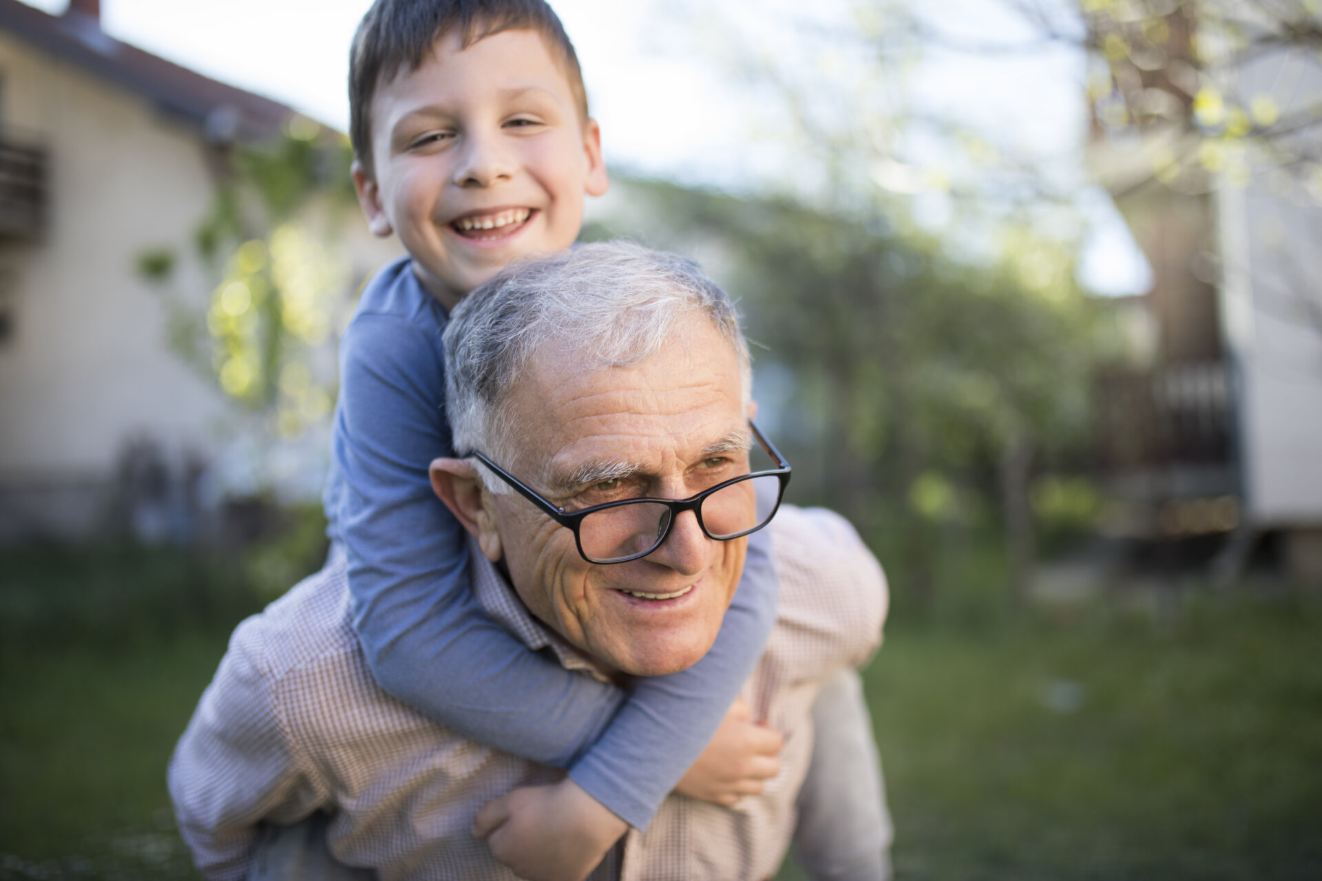 Ένας παππούς με το εγγόνι του, το οποίο έχει στους ώμους του και φαίνονται να απολαμβάνουν χαρούμενες στιγμές στον κήπο του σπιτιού τους.