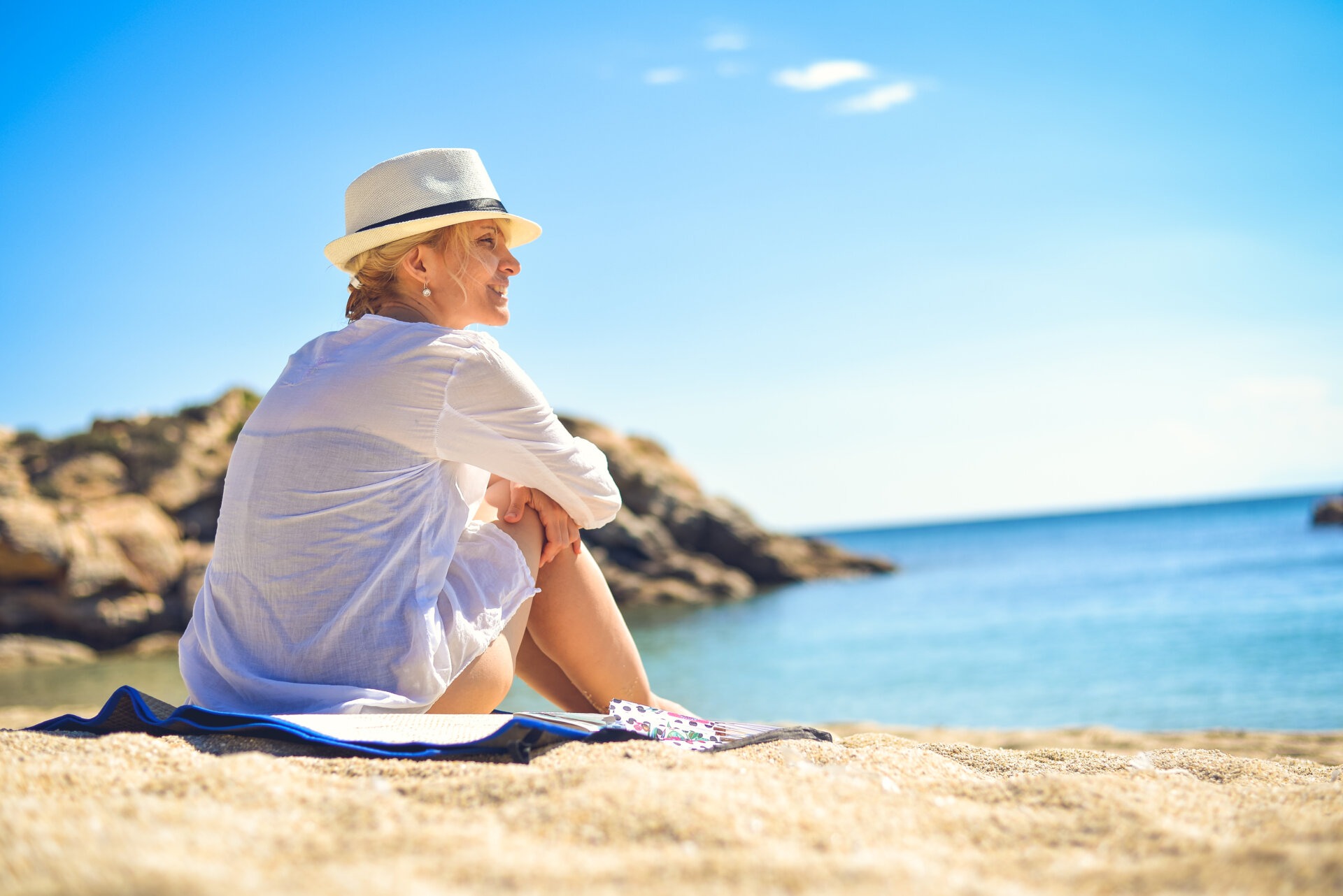 Μια γυναίκα, σε μια παραλία, που φοράει ένα καπέλο, ένα λινό πουκάμισο και το μαγιό της, και κάθεται στην άμμο, πάνω σε μια πετσέτα.