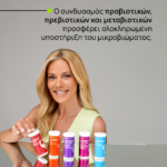 Γυναίκα με ξανθά μαλλιά χαμογελά μπροστά σε γκρι φόντο, τοποθετεί σωληνάκια προβιοτικών σε γυαλισμένο τραπέζι.