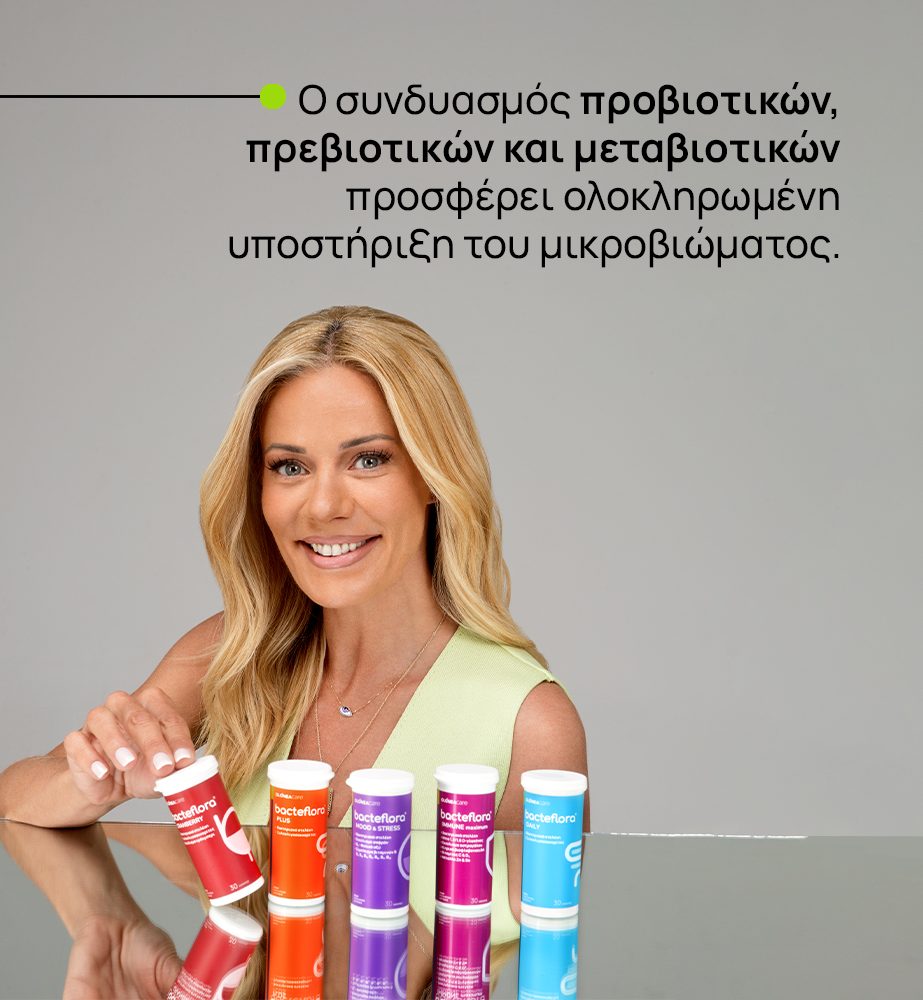 Γυναίκα με ξανθά μαλλιά χαμογελά μπροστά σε γκρι φόντο, τοποθετεί σωληνάκια προβιοτικών σε γυαλισμένο τραπέζι.