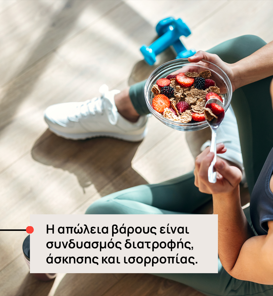 Άτομο σε γυμναστήριο/σπίτι, τρώει μπολ δημητριακών με φρούτα (φράουλες, μύρτιλα) ενώ υπάρχουν αλτήρες στο παρασκήνιο — μήνυμα ότι η απώλεια βάρους εξαρτάται από διατροφή, άσκηση και ισορροπία.